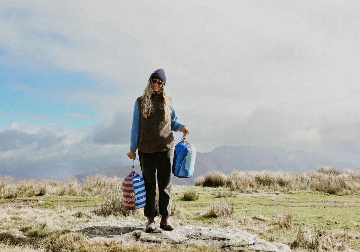 Smiling woman on a hilltop holding two packed VOITED Cloudtouch blankets in their stuff sacks.