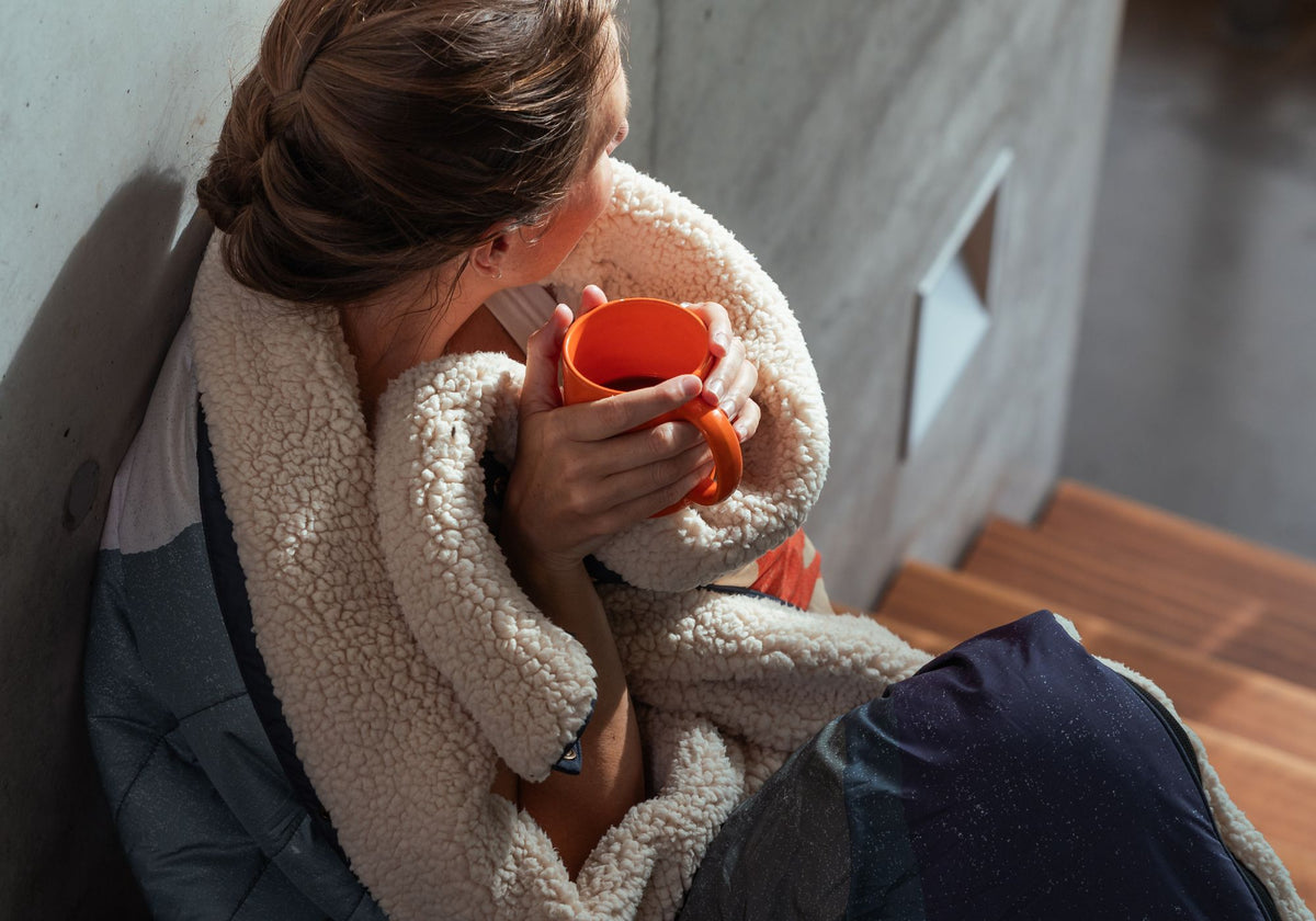 A woman sits on a wooden bench wrapped in a cream sherpa fleece Cloudtouch blanket, holding an orange mug. She is wearing a gray quilted puffer jacket and a white top, set against a concrete wall and wooden stairs.VOITED