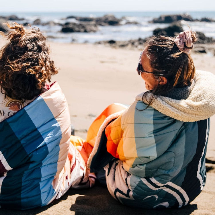 Two people sitting on a sandy beach facing the ocean, wrapped in colorful Cloudtouch blankets VOITED with blue, orange, cream, and gray panels.
