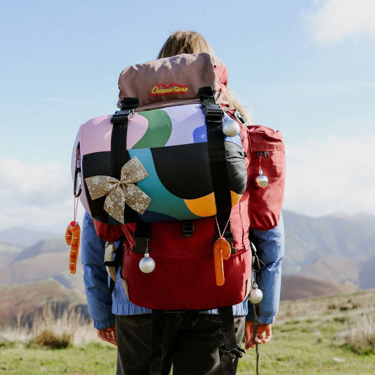 A person viewed from behind wearing a large, highly decorated hiking backpack on a mountain trail. Cloudtouch VOITED Blanket.