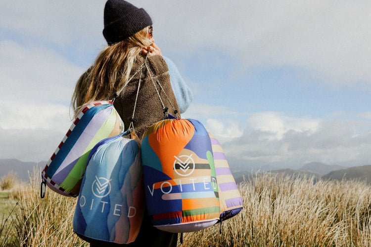 Woman carrying four colorful VOITED outdoor blankets in stuff sacks over her shoulder, with grassland and mountains behind.