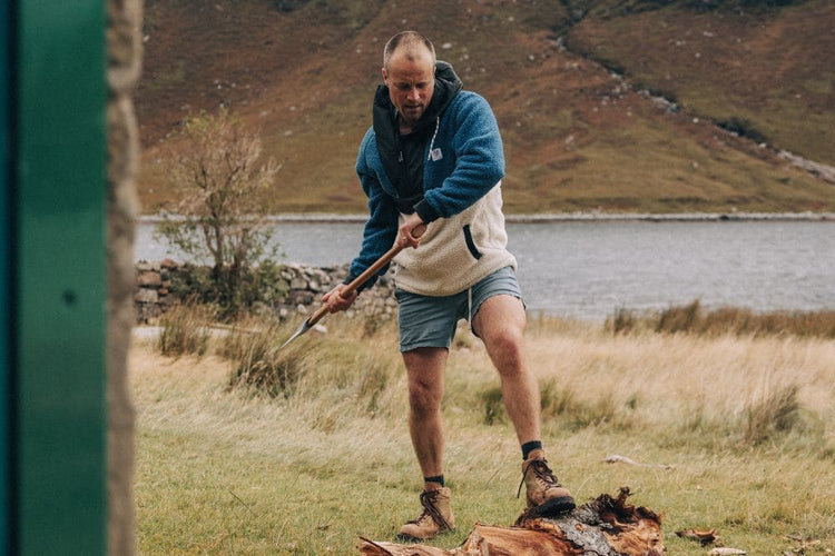 Man in outdoor clothing using a tool by a campfire with a scenic lake and mountains in the background